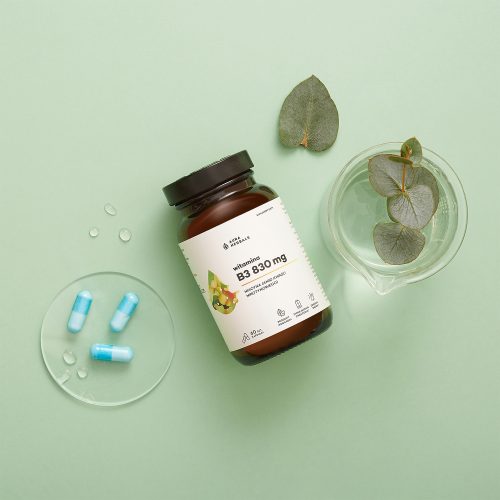 Butelka z napisem "Witamina B3 830 mg, niacyna, kapsułki 60 szt" spoczywa na zielonej powierzchni obok dwóch niebieskich kapsułek, szklanego naczynia z kroplami wody i liści eukaliptusa.