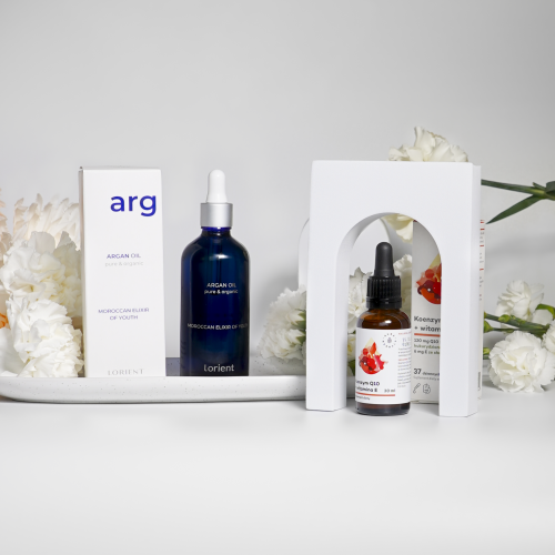 Dwa produkty do pielęgnacji skóry aura herbals - zestaw 6-6, wraz z ich pudełkami, są wyświetlane na białej powierzchni wśród białych kwiatów. Jeden z nich zawiera olej arganowy, a drugi koenzym Q10.