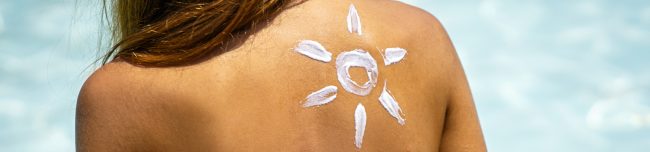 Dziewczyna nakłada na skórę balsam do opalania Skin and Body Care i nawilżający filtr przeciwsłoneczny, a na jej plecach narysowany jest kształt słońca na rozmytym niebieskim tle.