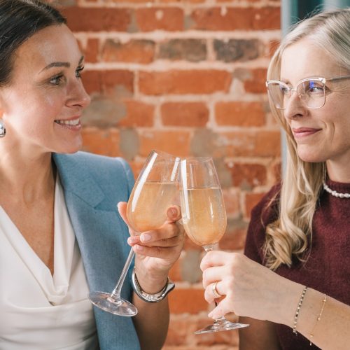 Dwie kobiety uśmiechają się i brzęczą kieliszkami, siedząc przed ceglaną ścianą, ciesząc się Zestaw świąteczny Kolagen + Srebro Argentum200_4_1500x1500.