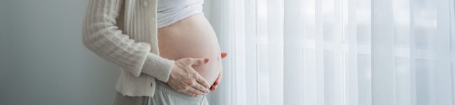 Kobieta w ciąży stoi w pomieszczeniu w pobliżu okna z przezroczystymi zasłonami, delikatnie trzymając obiema rękami swój nagi brzuch.