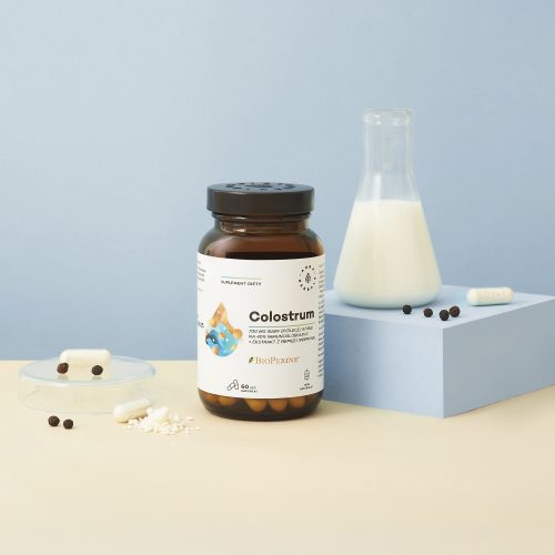 Butelka z napisem „colostrum-700-mg-bioperine-kapsulki-60-szt_2_1500x1500” stoi obok szklanej kolby z mlekiem, kapsułkami i czarnymi kulkami na jasnoniebieskiej powierzchni na jasnoniebieskim tle.