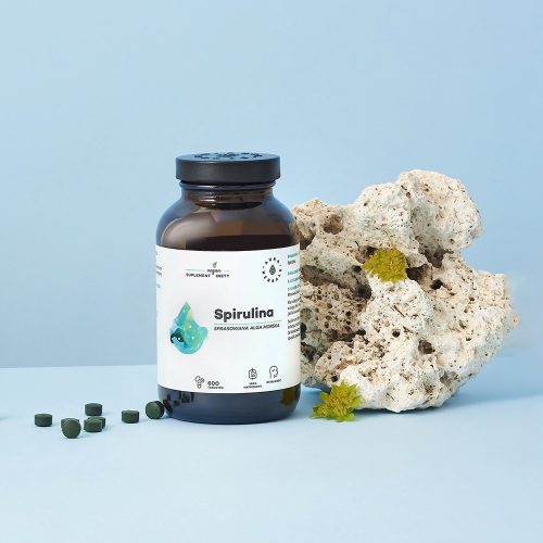 Butelka Spirulina-tabletki-600-szt.-1500x1500-2-1 stoi obok porowatej skały i małych kawałków zielonych glonów, a wszystko to na jasnoniebieskim tle.