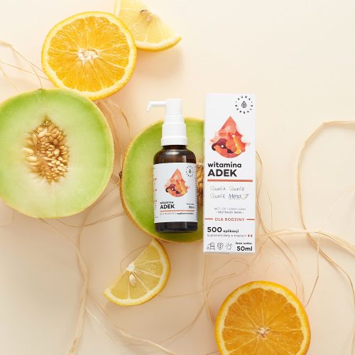 Na jasnym tle widoczna jest butelka i opakowanie produktu „Witamina-ADEK-dla-rodziny-MCT-krople-50-ml” z dozownikiem w postaci pompki, obok połowy melona, pokrojonej cytryny oraz ozdobnej słomki.