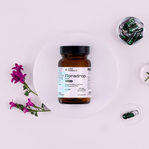 Brązowa szklana butelka z napisem „Floradrop-femi-probiotyk-kapsułki-20-szt.” stoi na białym talerzu z fioletowymi kwiatami, a obok niej leżą zielone kapsułki; całość na jasnym tle.