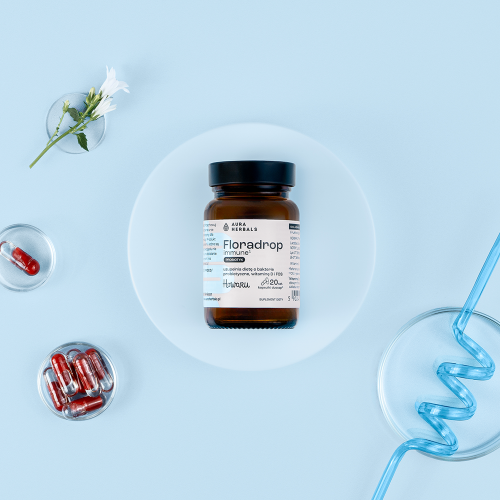 Na białym talerzu stoi brązowa buteleczka z napisem „Floradrop – immune – probiotyk – kapsułki – 20 szt.”, otoczona czerwonymi kapsułkami, białym kwiatem w szklance oraz niebieską słomką na jasnoniebieskim tle.