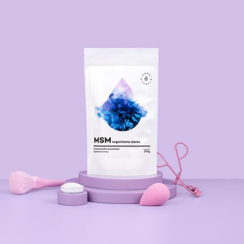 Na fioletowej podstawce stoi saszetka z produktem „MSM-Organiczny-Związek-Siarki-proszek-200-g”, otoczona różowymi i fioletowymi akcesoriami do makijażu, na lawendowym tle.