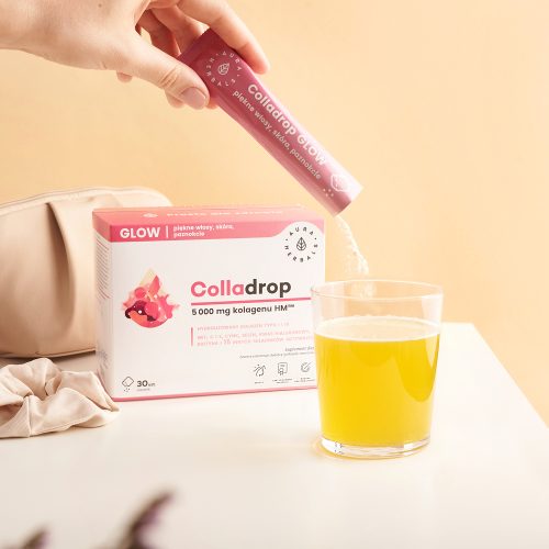 Ręka wsypuje proszek „Colladrop-Glow-kolagen-morski-5000-mg-proszek-saszetki-30-szt.” z różowej saszetki do szklanki z żółtym płynem, a na jasnym tle widoczne są opakowanie produktu oraz beżowa torebka.