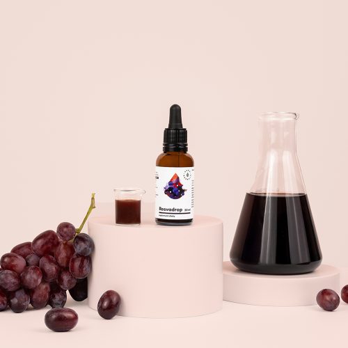 Na pastelowych podstawkach na jasnoróżowym tle ustawiono butelkę preparatu Resvadrop-Resweratrol (krople, 30 ml), małą szklankę z płynem, zlewkę zawierającą ciemny płyn oraz świeże winogrona.