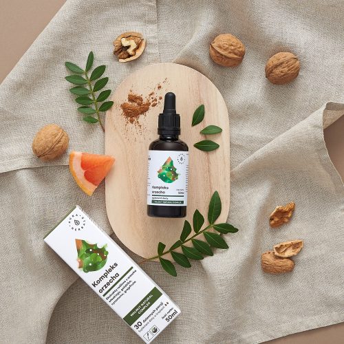 Na drewnianej desce, na tle beżowego materiału, widnieje buteleczka z zakraplaczem o pojemności 50 ml wraz z opakowaniem z napisem „Orzech-kompleks-krople-Walnut-Natural-Complex-50ml”, a obok niej orzechy włoskie, łupiny orzechów włoskich, zielone liście oraz kawałek grejpfruta.