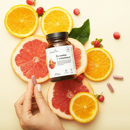 Na żółtym tle widać dłoń trzymającą słoik z suplementem „Zurawina – 800 mg – witamina C – kapsułki – 60 szt.”, otoczony plasterkami pomarańczy, grejpfrutów, żurawiny i zielonych liści.