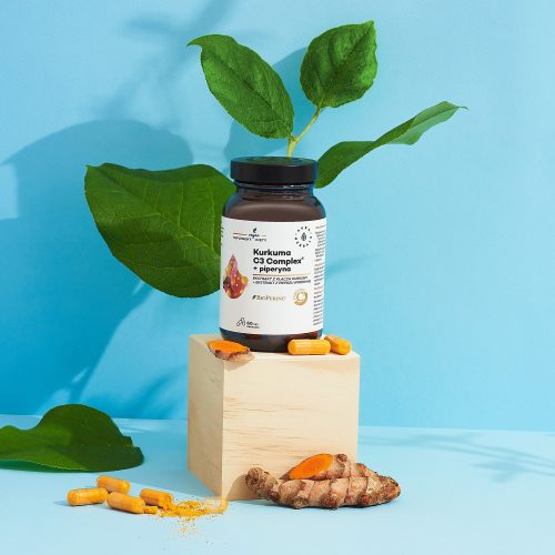 Na drewnianej podstawce, na niebieskim tle, prezentowana jest butelka kapsułek z suplementem „kurkuma-500-mg-c3-complex-piperyna-kapsulki-60-szt_2_1500x1500” wraz z korzeniem kurkumy, luźnymi kapsułkami i zielonymi liśćmi.