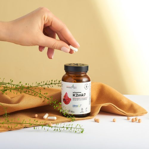 W dłoni trzyma się białą kapsułkę nad brązową butelką z napisem „witamina-k2-menaq7-200-ug-vegan-kapsulki-90-szt-_1500x1500-scaled-1”. Na stole leżą porozrzucane tabletki, łodyga rośliny i pomarańczowa ściereczka.