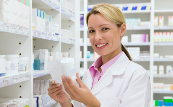 Farmaceuta w białym fartuchu laboratoryjnym stoi przed półkami w aptece, trzymając w ręku suplementy diety wskazane przy osteoporozie